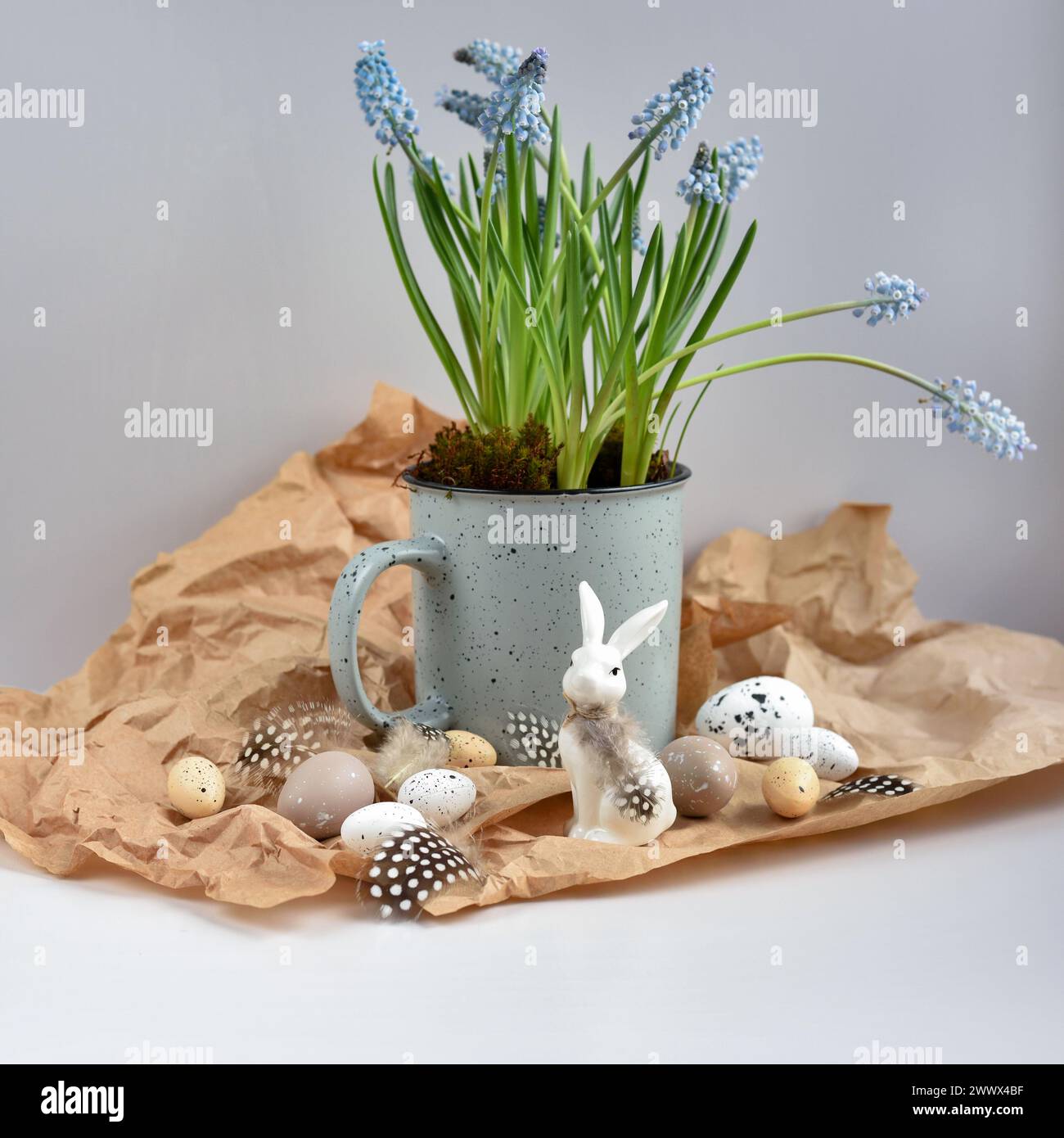 Osterkomposition mit weißem Kaninchen, Eiern, Federn und hellblauen Muscari-Blüten in Tasse. Osterstillleben Stockfoto