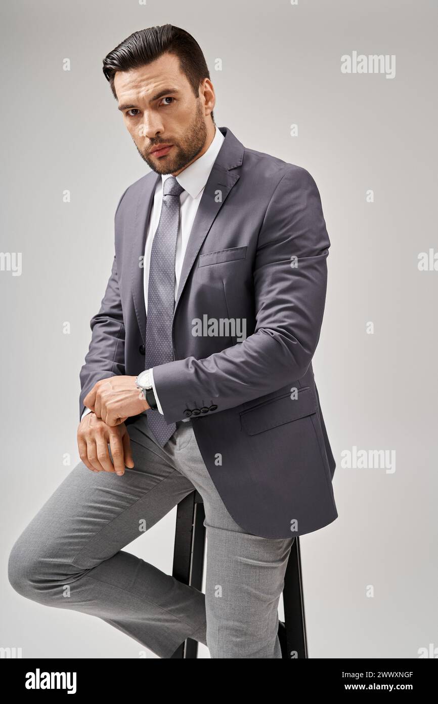Gutaussehender und selbstbewusster Geschäftsmann mit Borsten, der im Anzug posiert und auf einem hohen Hocker auf Grau sitzt Stockfoto