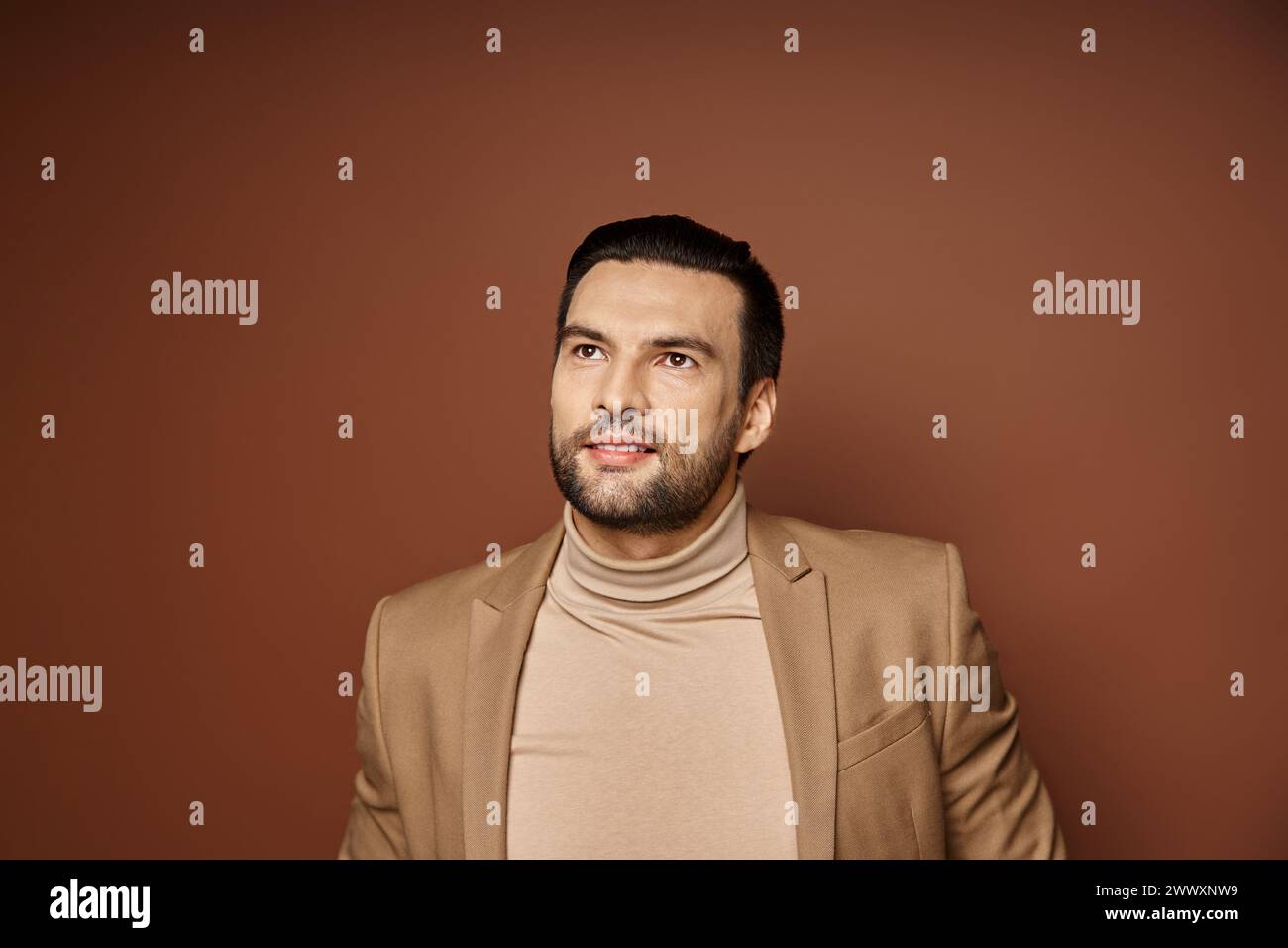 Traumhafter und gutaussehender Mann mit lächelnden Borsten und posiert in stilvollem beigefarbenem Blazer vor beigefarbenem Hintergrund Stockfoto
