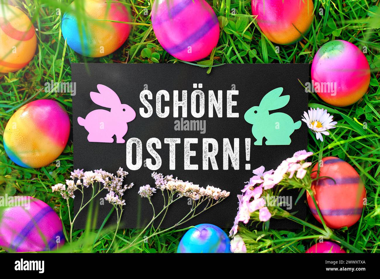 26. März 2024: Ostergrüße: Frohe Ostern auf einem Schild auf einer Wiese mit bunten Ostereiern. FOTOMONTAGE *** Ostergruß: Schöne Ostern auf einem Schild in einer Wiese mit bunten Ostereiern. FOTOMONTAGE Stockfoto