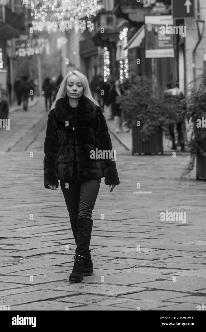 Schwarz-weiß-Bild einer Frau, die tagsüber in einem Pelzmantel auf einer Stadtstraße läuft Stockfoto