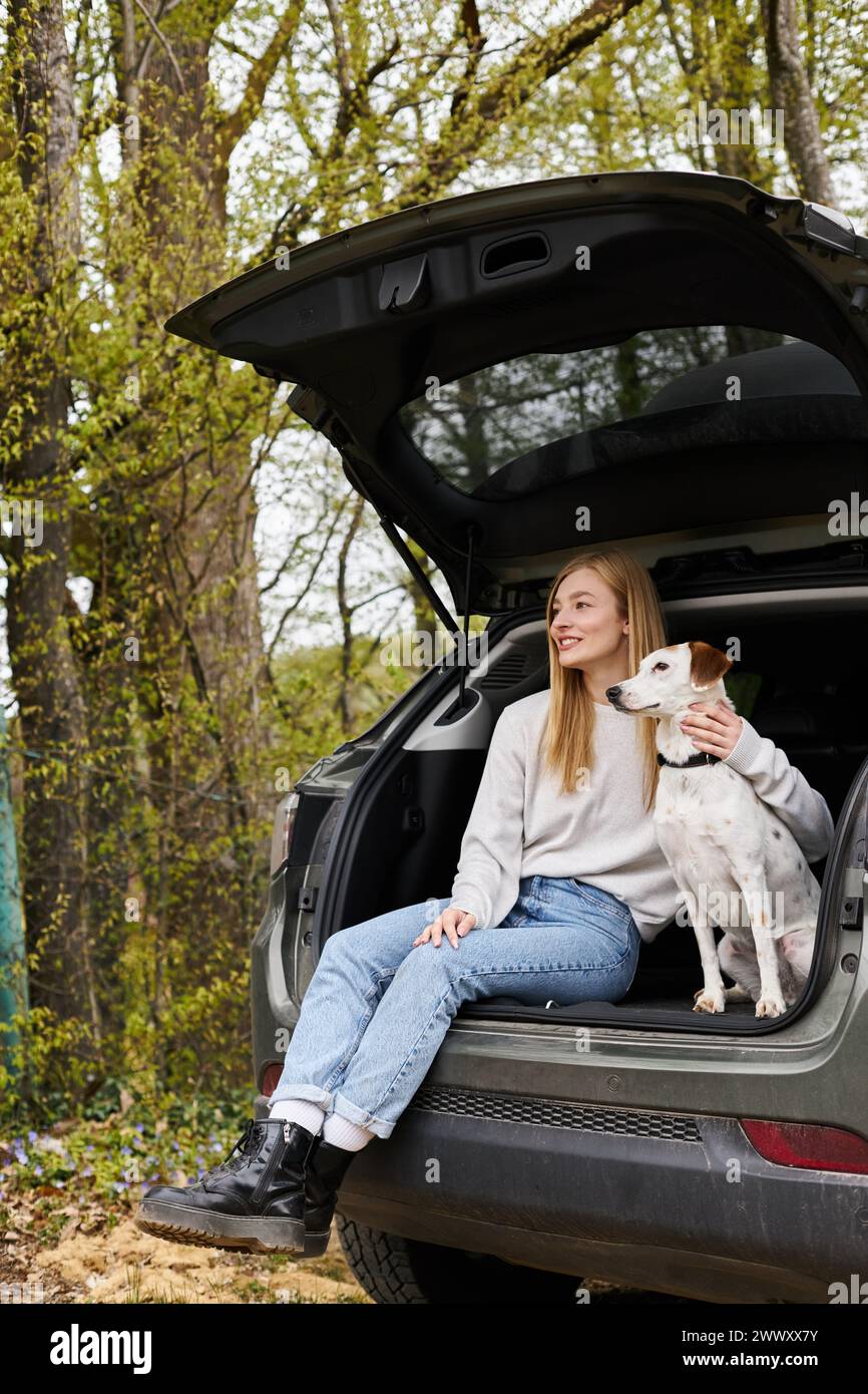 Entspannte Frau, die ihren Hund umarmt, im Auto sitzt und im Wald wegblickt, um zu wandern Stockfoto