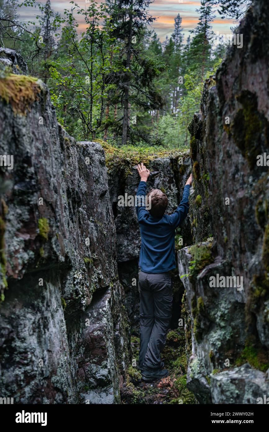 Junger Entdecker, der eine Waldspalte in Dalarna, Schweden, skaliert. Junge klettert zwischen moosbedeckten Felswänden unter dem schwedischen Sommerabendhimmel. Stockfoto