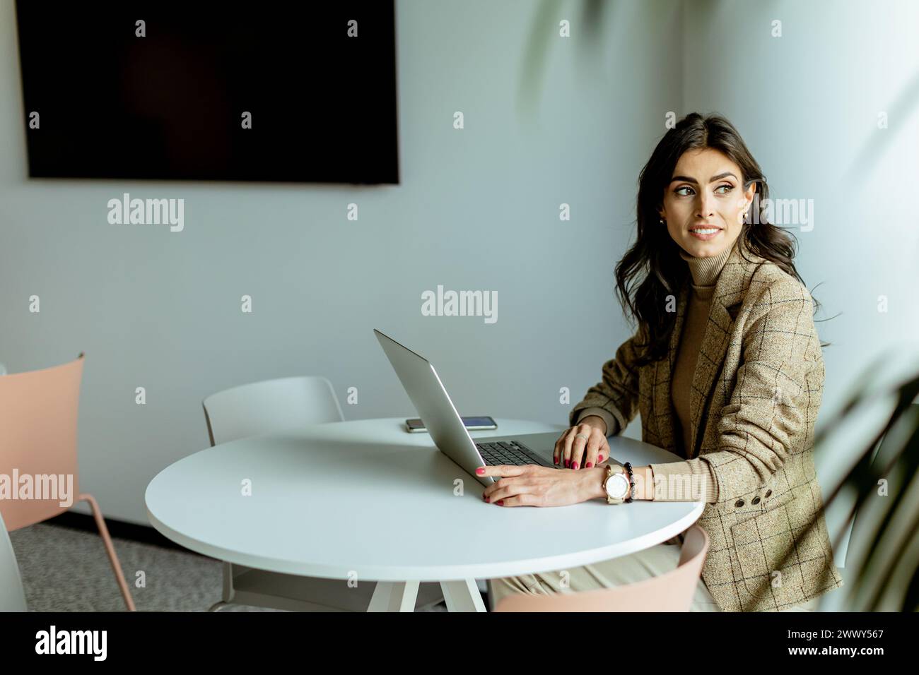 Eine konzentrierte Geschäftsfrau in einem stilvollen Blazer lässt sich auf ihrem Laptop an einem eleganten, weißen Tisch, umgeben von minimalistischem Dekor, verstecken. Stockfoto