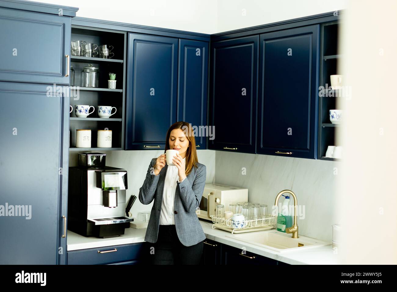 Eine lächelnde Frau in Geschäftskleidung, die eine Kaffeetasse hält und in einer stilvollen blauen Küche eine entspannende Pause einlegt. Stockfoto