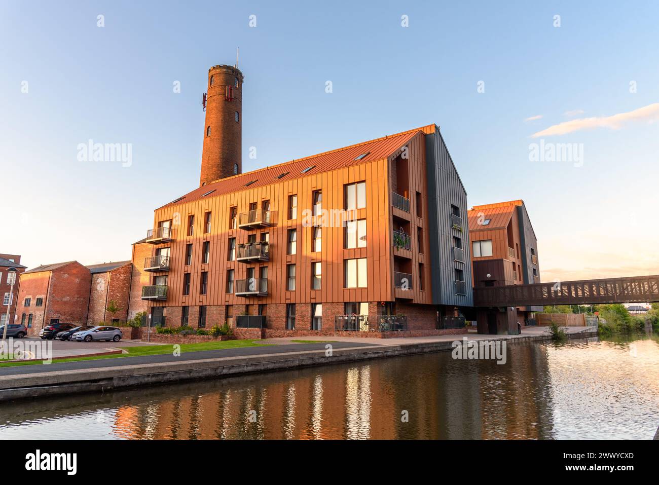 Neue Apartmentgebäude mit Blick auf einen historischen Backsteinturm entlang eines Kanals unter klarem Himmel bei Sonnenuntergang Stockfoto