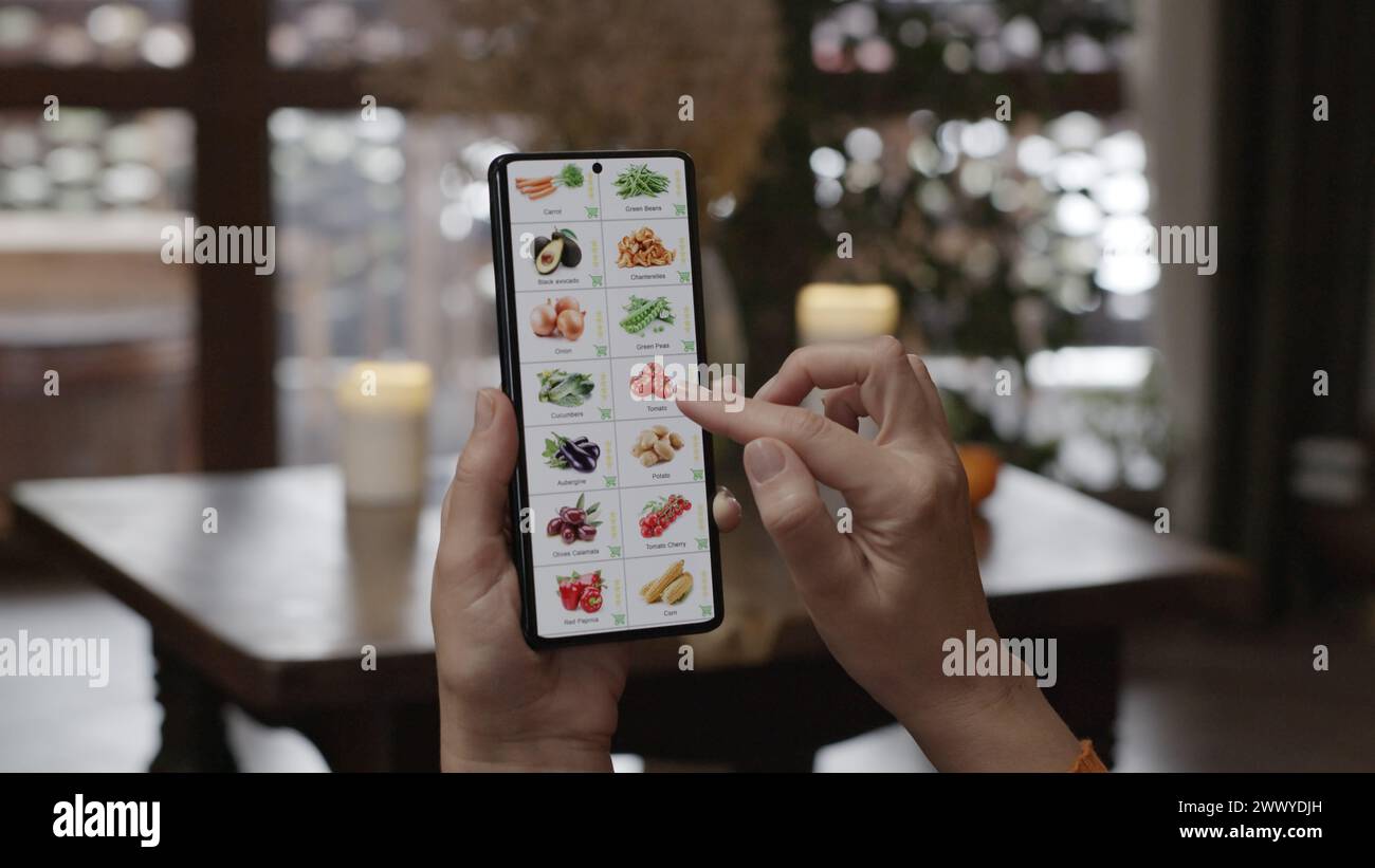 Lebensmittel mit einem Smartphone zu Hause bestellen. Eine Frau wählt Gemüse in einem Online-Shop mit einer Anwendung auf einem Smartphone aus. Heim-Abendmöbel Stockfoto