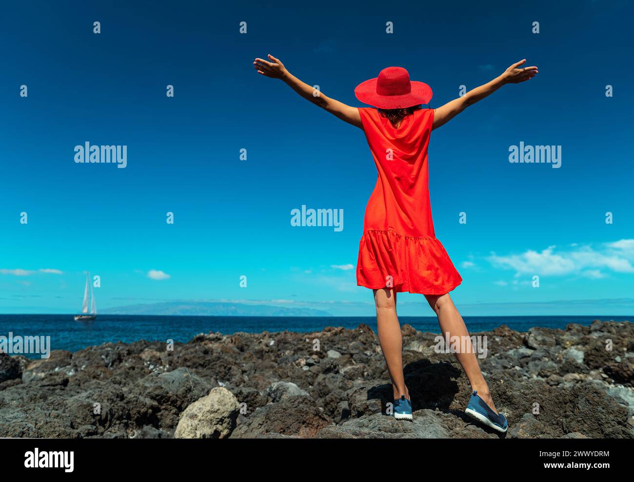 Glückliche Frau in Rot, die mit offenen Händen am felsigen Meer steht. Ruhiges Ozeanwasser und blauer Himmel im Hintergrund. Stockfoto