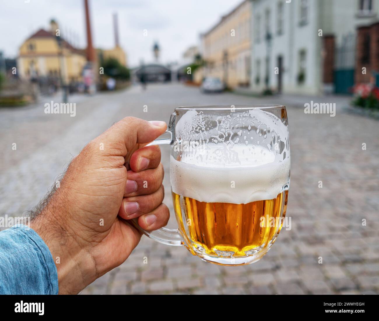 Ein Mann hält einen Becher Bier in der Hand und verschwommene Pflastersteine und altmodische Häuser im Hintergrund. Stockfoto