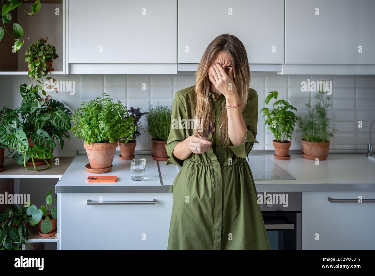 Unwohl müde Frau, die Gläser abzieht, die Augen mit der Hand auf der Küche zu Hause mit Zimmerpflanzen reibt. Stockfoto