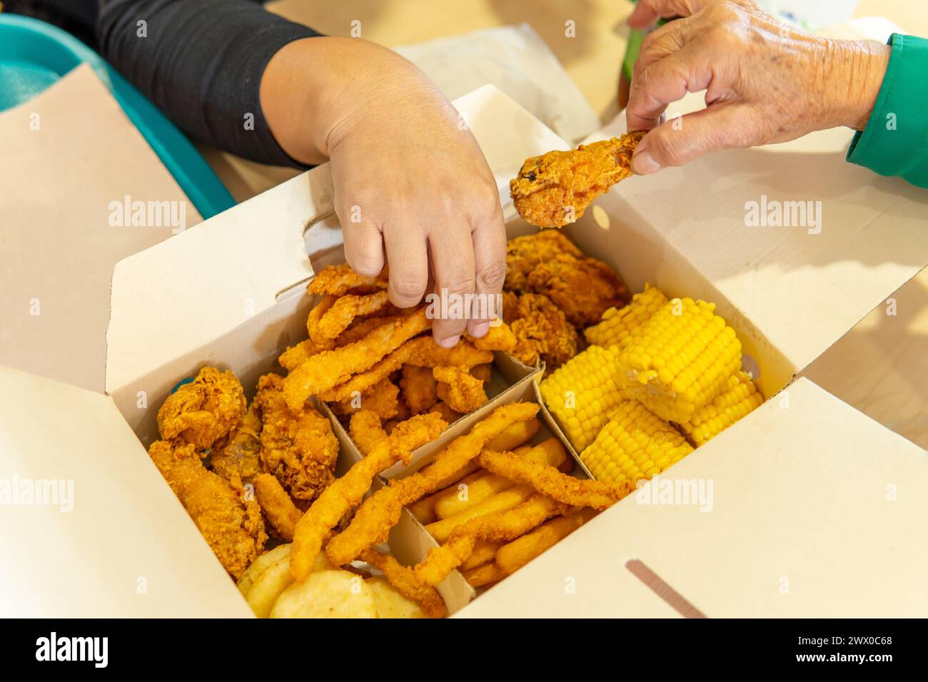 Hände von zwei Frauen nehmen Essen aus einer Fast-Food-Box mit Hühnchen, Mais, Pommes Frites und Arepas in einem Food Court Stockfoto