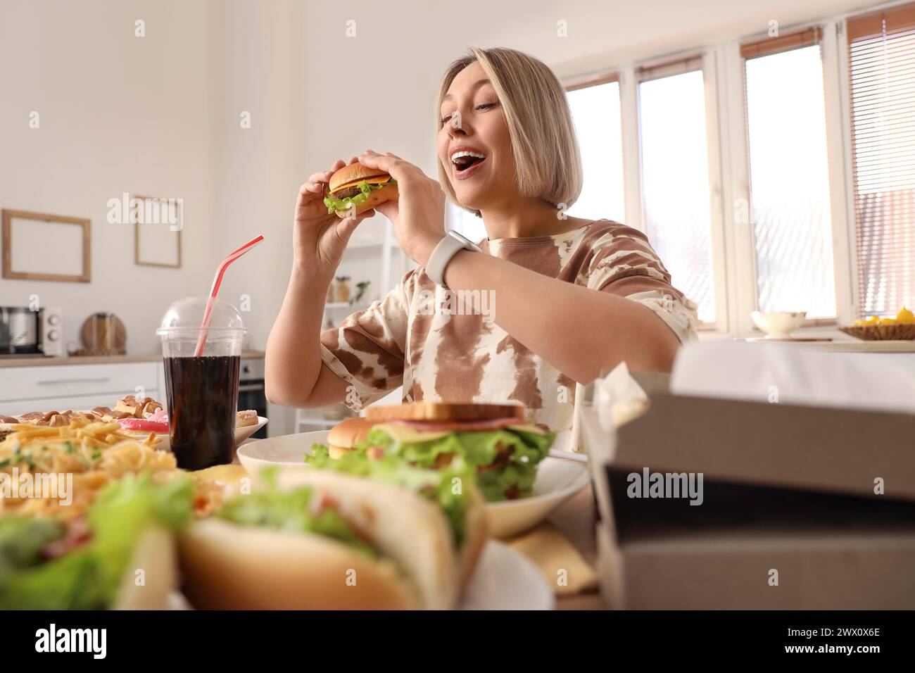 Schöne Frau mit leckerem Burger am Tisch in der Küche. Das Konzept des Überessens Stockfoto