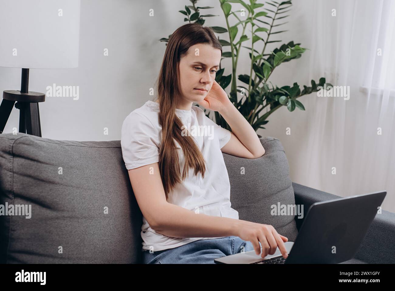 Fokussierte freischaffende Mädchenarbeit am Laptop auf gemütlicher Couch in modernem Wohnzimmer, sieht durchdachte Such-Ideen aus, korrespondiert mit dem Kunden, löst Bus Stockfoto