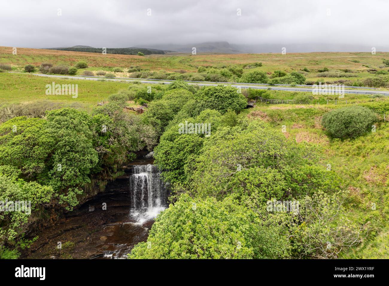 Eingebettet in üppiges Grün, bilden die Lealt Falls eine Oase der Ruhe auf Skye, ergänzt durch eine Landstraße und das entfernte, nebelbehüllte Hochland Stockfoto