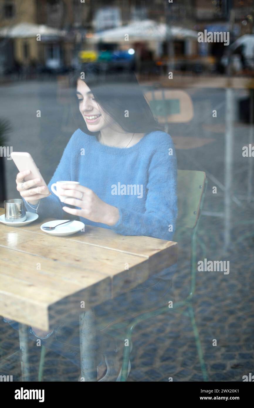 Eine junge Frau in einem blauen Pullover fokussiert sich in einem Café auf ihr Handy, mit einer Kaffeetasse und Untertasse vor ihr, durch ein Fenster gesehen Stockfoto