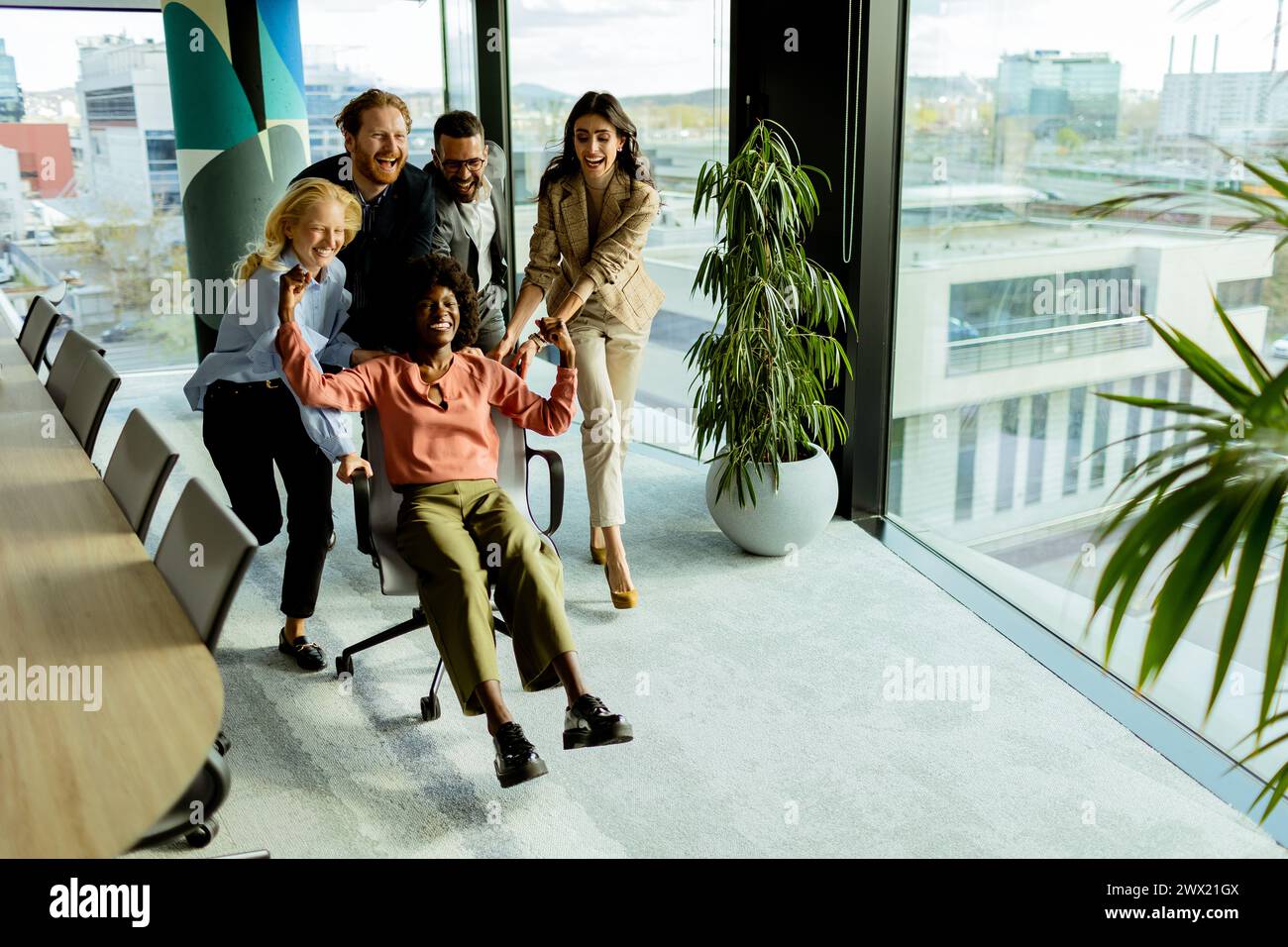 Ein Team von Kollegen teilt sich einen Moment des Spaßes und fährt mit einem Bürostuhl inmitten eines gut beleuchteten Arbeitsplatzes. Stockfoto