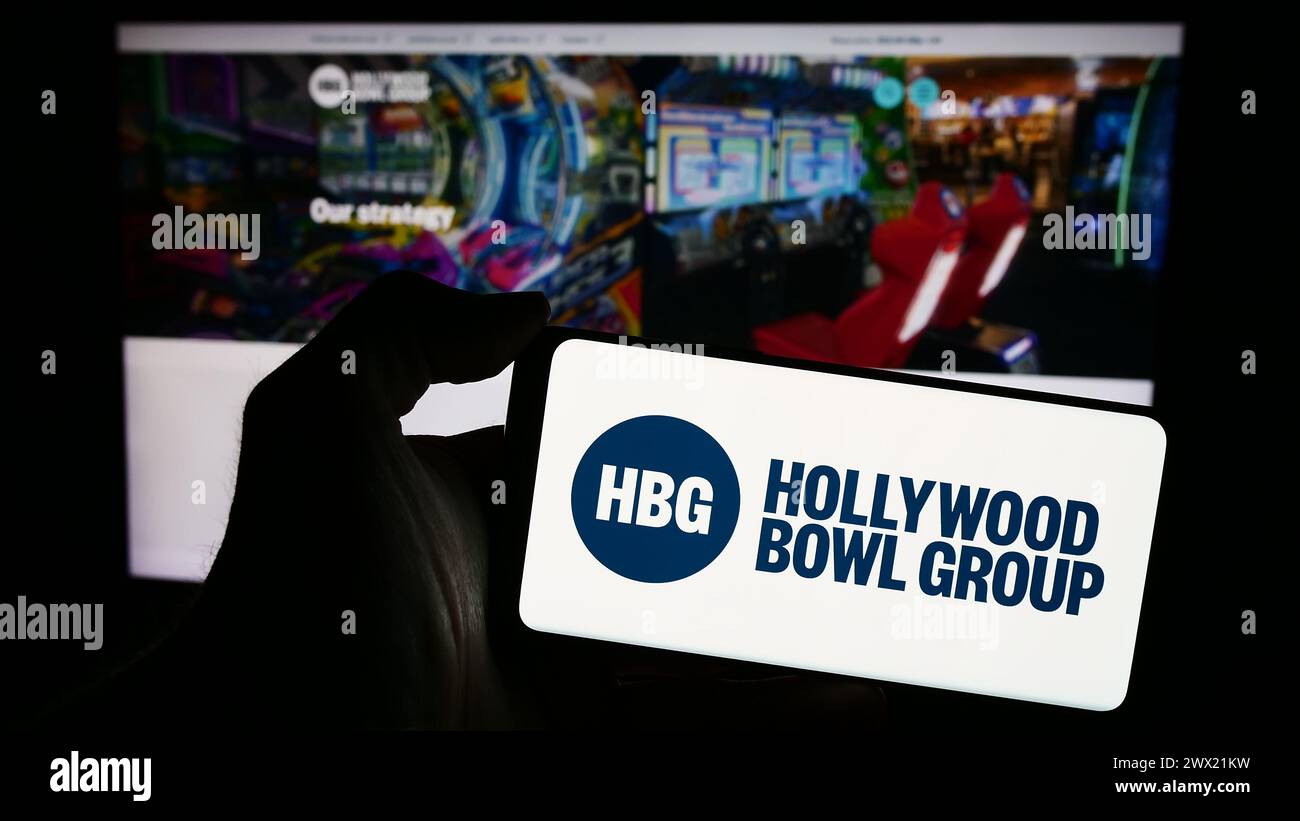 Person mit Smartphone und Logo der britischen Bowlingfirma Hollywood Bowl Group plc vor der Website. Schwerpunkt auf dem Display des Telefons. Stockfoto