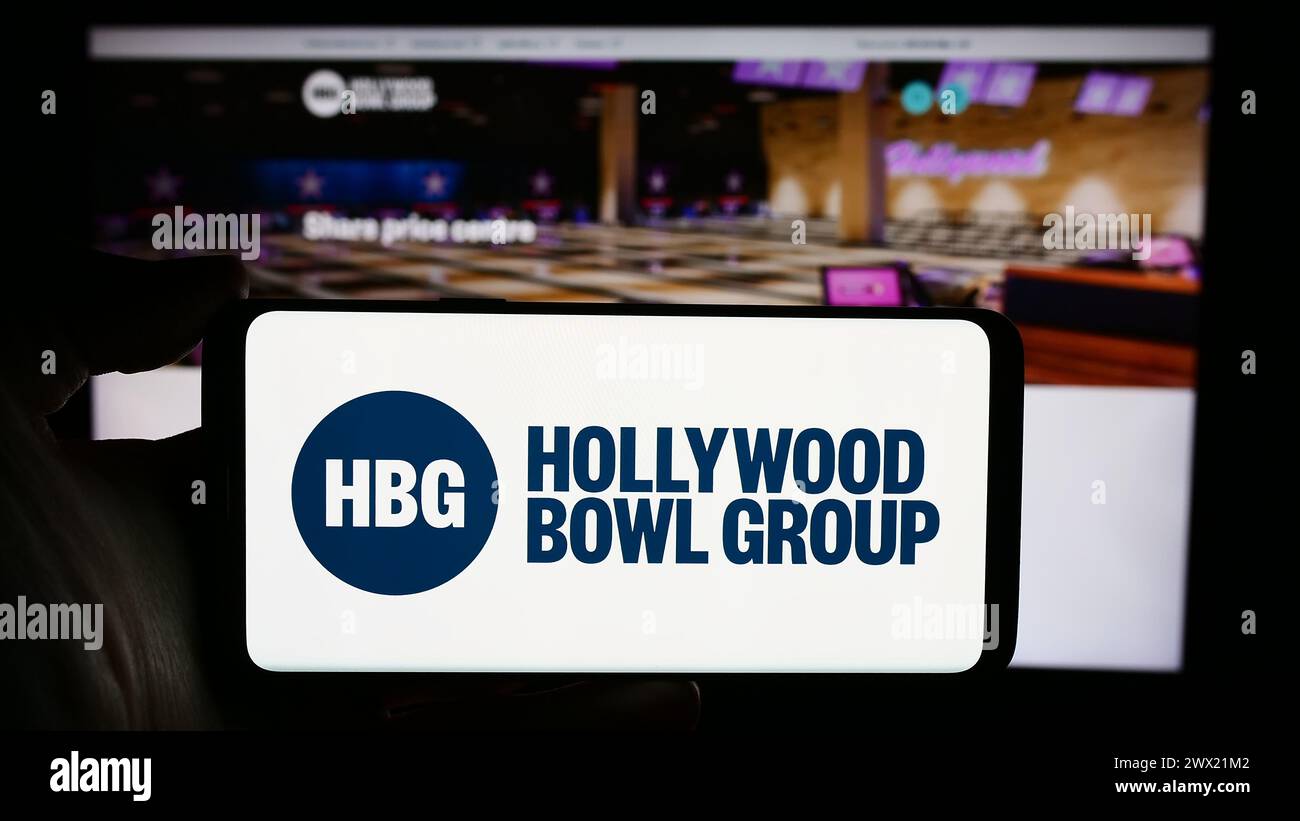 Person mit Handy mit Logo der britischen Bowlingfirma Hollywood Bowl Group plc vor der Business-Website. Schwerpunkt auf dem Display des Telefons. Stockfoto