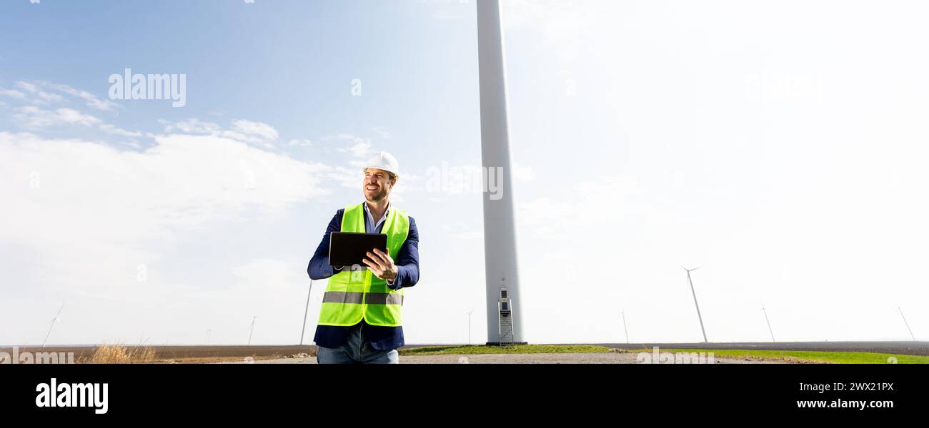 Ein Ingenieur mit Sicherheitshelm und Klemmbrett untersucht die Funktionsweise einer majestätischen Windturbine inmitten üppiger Grünflächen unter klarem Himmel. Stockfoto