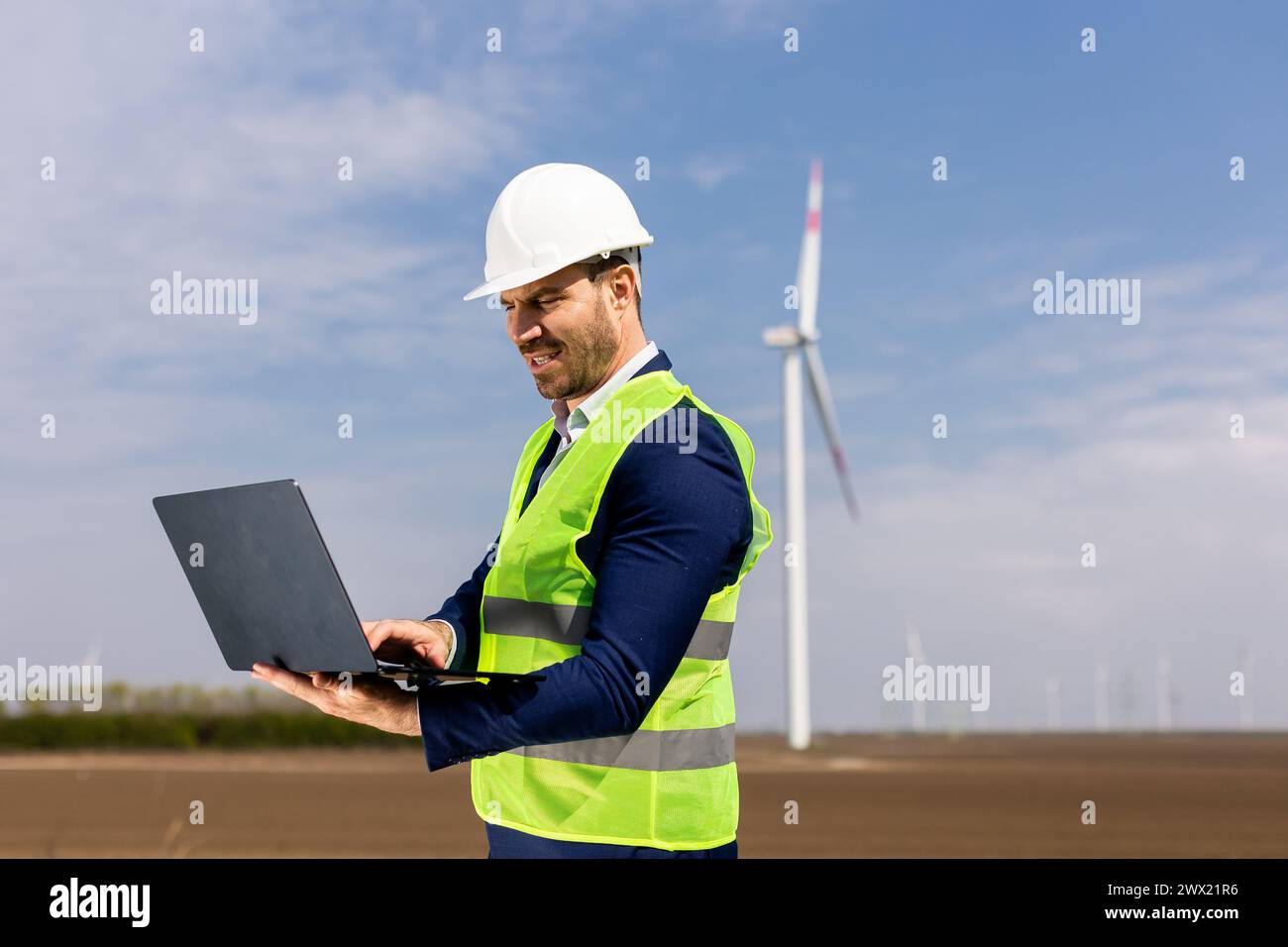 Ein fokussierter Ingenieur untersucht seinen Laptop mit einer Windturbine im Hintergrund unter klarem Himmel. Stockfoto