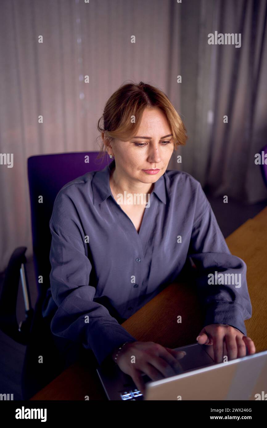 Eine schöne Geschäftsfrau mittleren Alters, die ein graues Hemd trägt und einen Laptop benutzt, während sie an einem modernen Arbeitsplatz sitzt Stockfoto