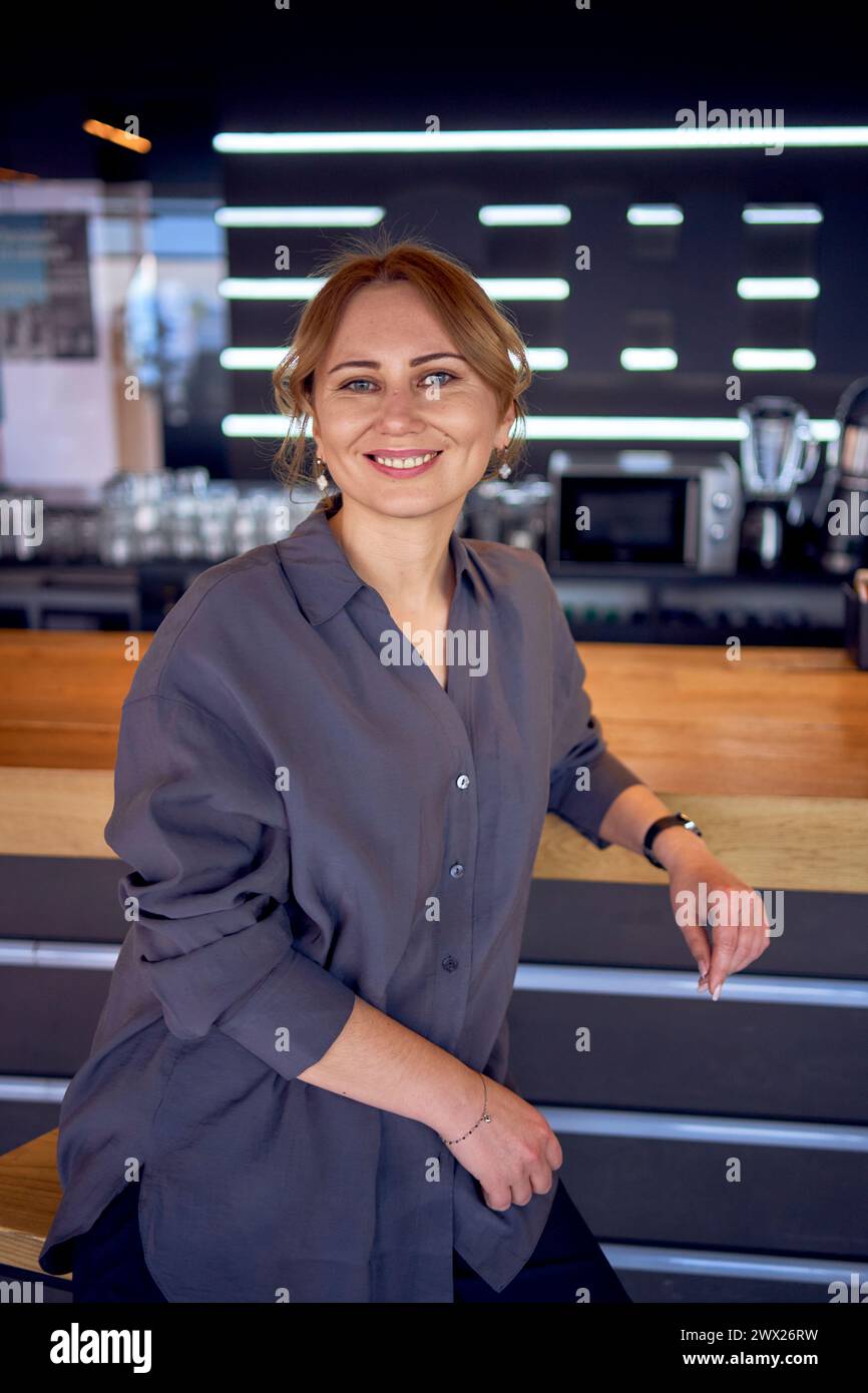 Eine attraktive Frau mittleren Alters in einer Bar mit neutralem Design, in einer Hose mit weitem Bein und einer Seidenbluse Stockfoto