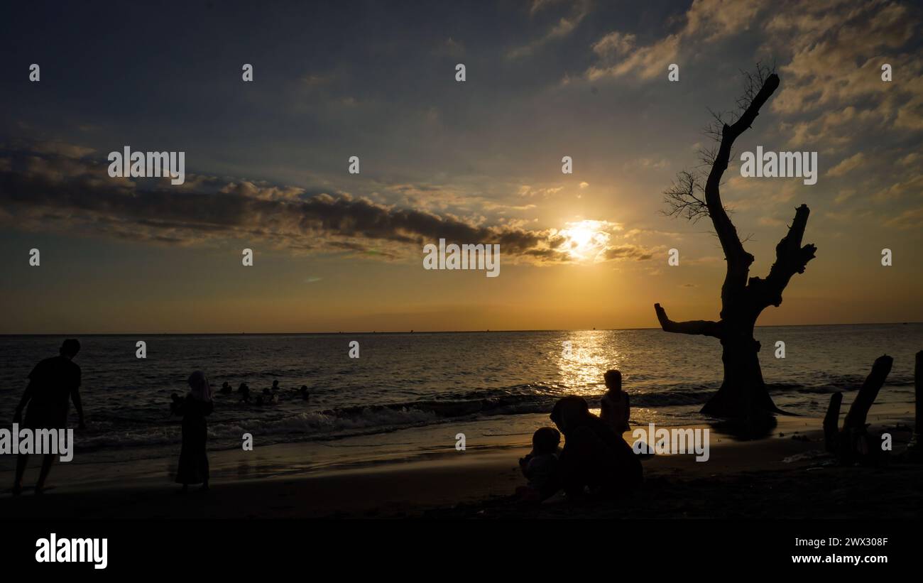 Die Sonne untergeht am Nachmittag am Ampenan Strand in Lombok mit der Silhouette von Ästen ohne Blätter und die Leute, die sich am Strand entspannen Stockfoto