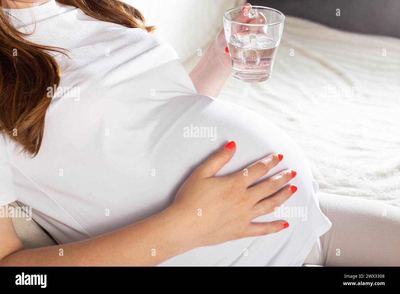 Ein schwangeres Mädchen mit einem Becher Wasser vor dem Hintergrund eines großen Bauches. Das Konzept von Durst und Wasserhaushalt bei Schwangeren. Trinkregime Stockfoto