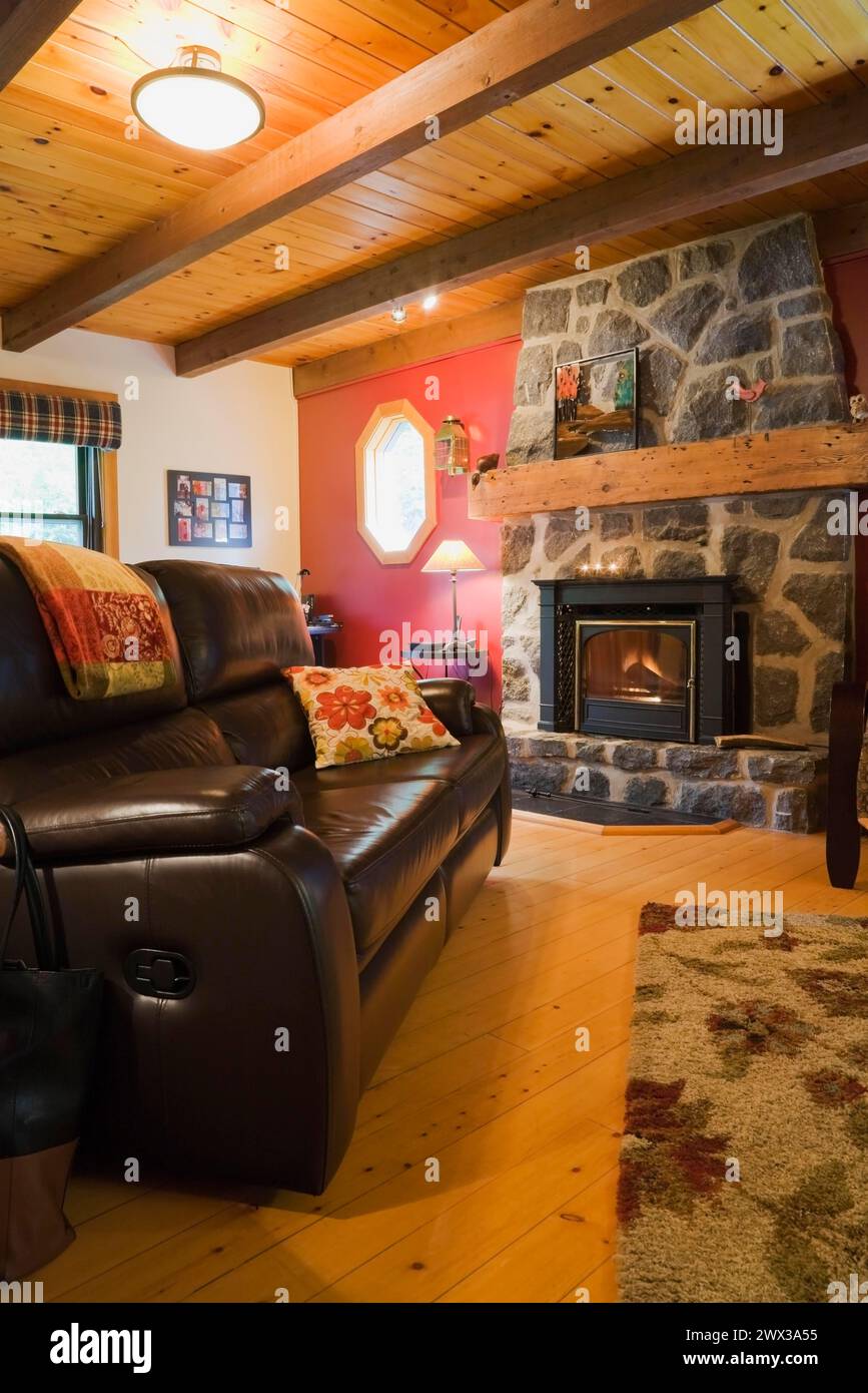 Beleuchteter Holzbrenner dunkler grauer Natursteinkamin und braunes Ledersofa im Wohnzimmer mit Hartholzboden im Blockhaus im kanadischen Stil Stockfoto