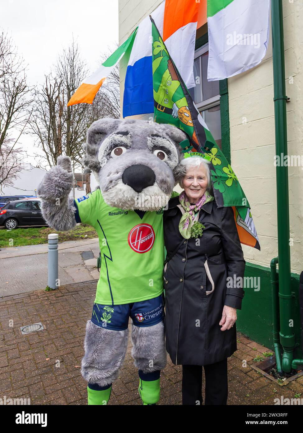 Die Dame trägt Happy St Patrick's Day Bonce Boppers und steht mit dem Warrington Rugby League Maskottchen Wolfie vor dem Warrington Irish Club Stockfoto