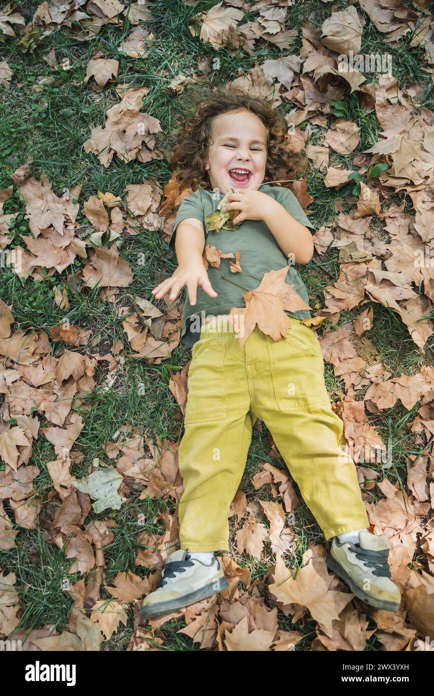 Glückliches Kind, das auf Gras mit trockenen Blättern spielt, Ganzkörperfoto Stockfoto