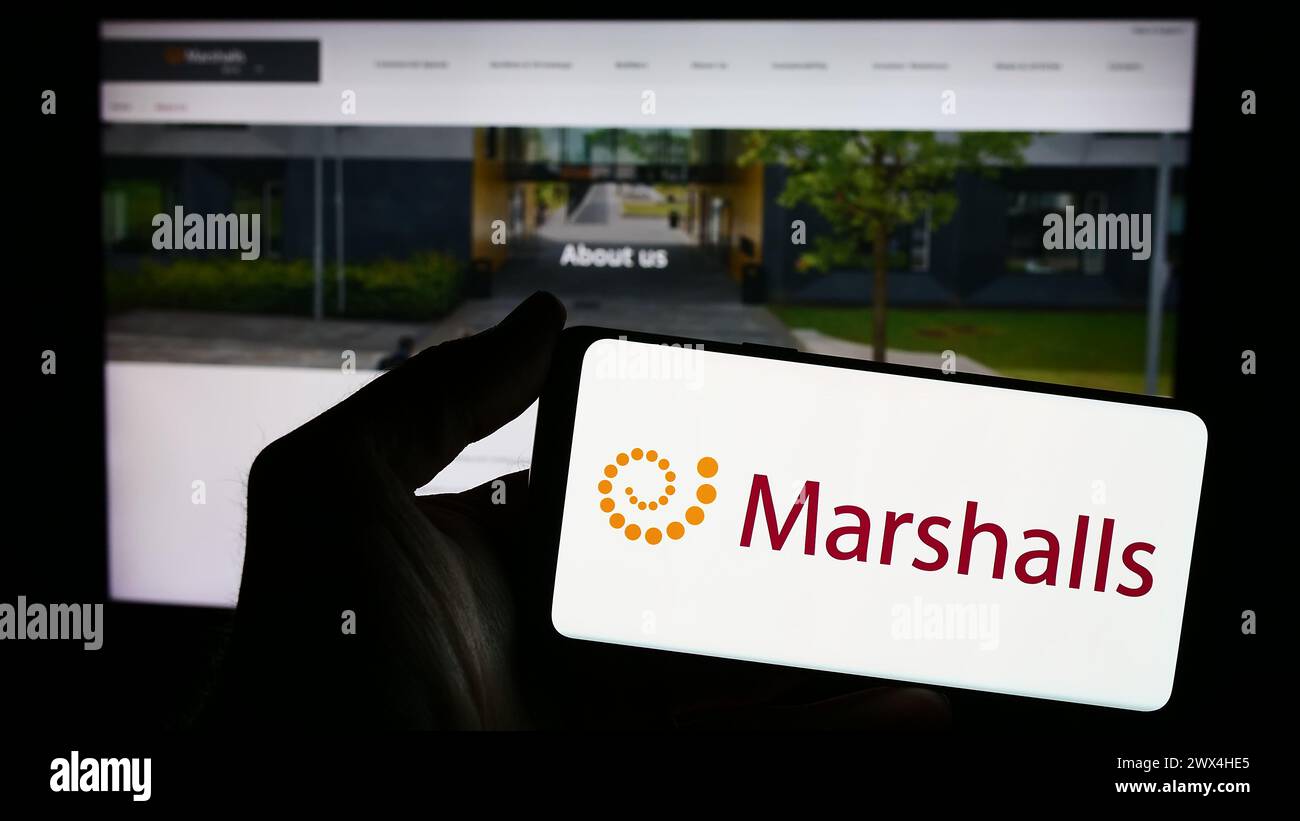 Person mit Smartphone und Logo des britischen Landschaftsprodukteunternehmens Marshalls plc vor der Website. Schwerpunkt auf dem Display des Telefons. Stockfoto