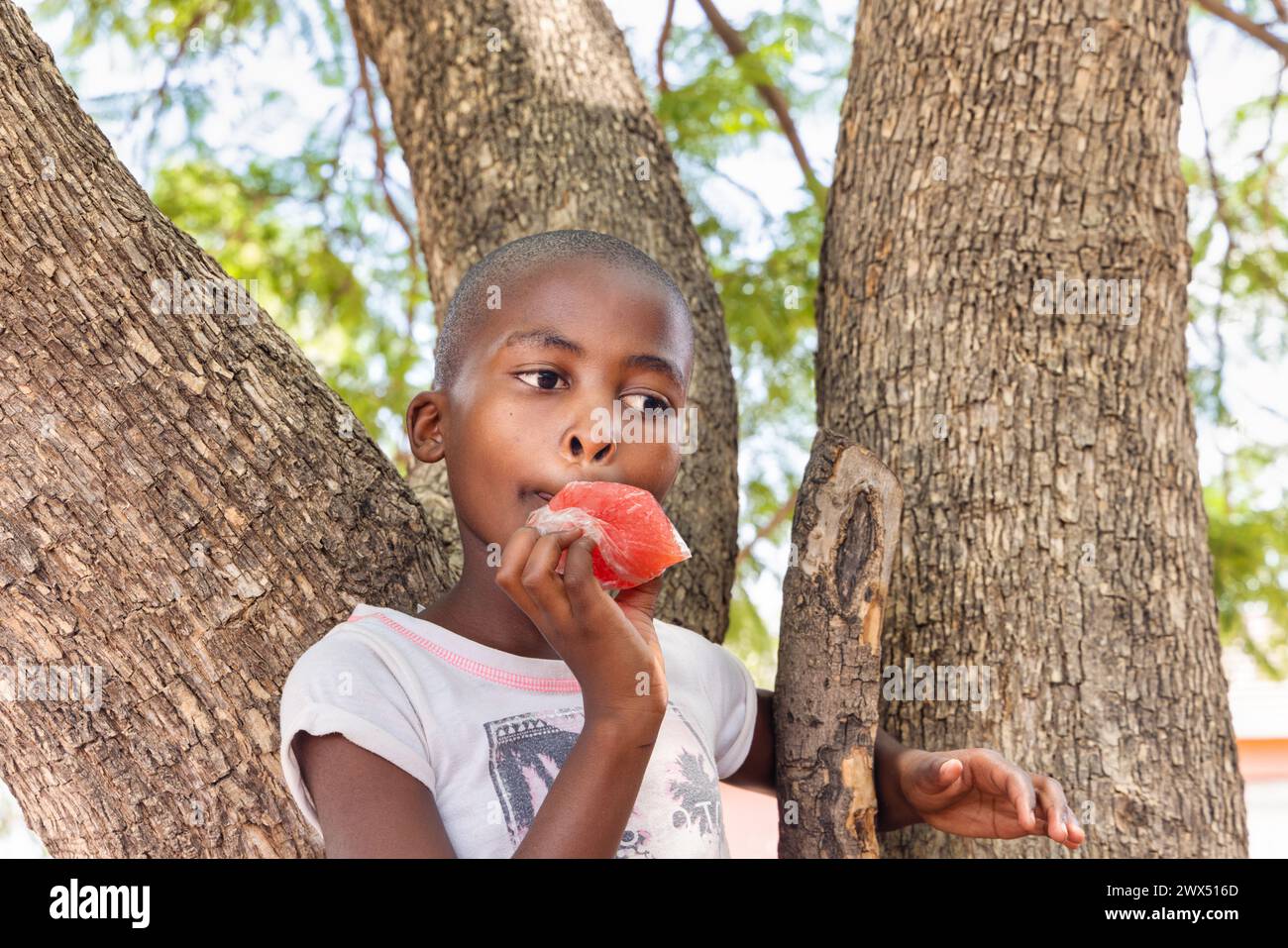 Porträt eines afrikanischen Dorfkindes, der gefrorenen Saft in einer Tasche isst, im Hof vor dem Haus, Dorfleben Stockfoto