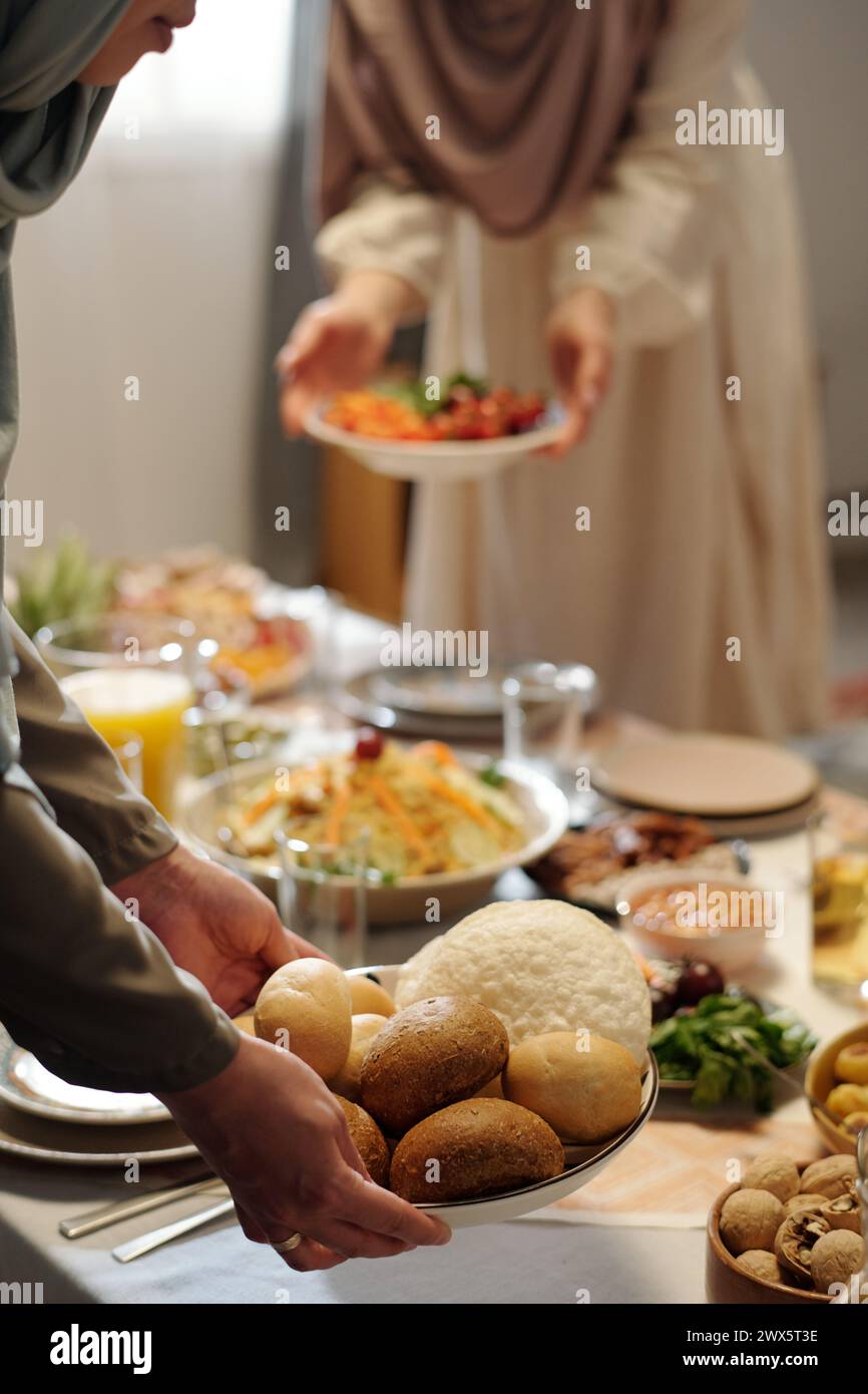 Vertikaler selektiver Fokus von zwei nicht erkennbaren muslimischen Frauen, die Hijabs tragen und einen festlichen Tisch für die Uraza Bayram-Feier legen Stockfoto