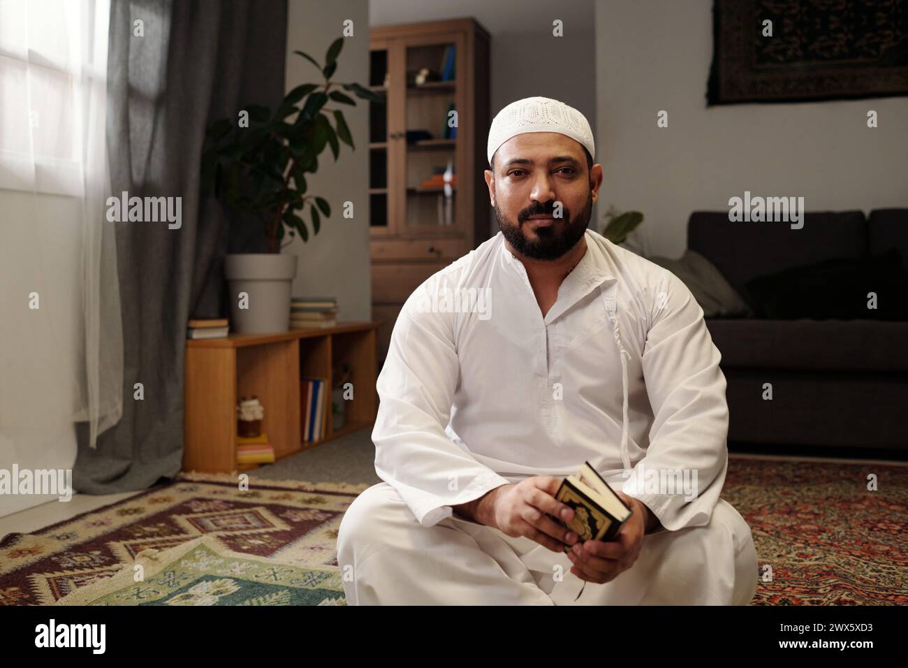 Porträt eines bärtigen muslimischen Mannes, der auf dem Boden im Wohnzimmer sitzt, mit Koranbuch in den Händen, Blick auf die Kamera, Kopierraum Stockfoto