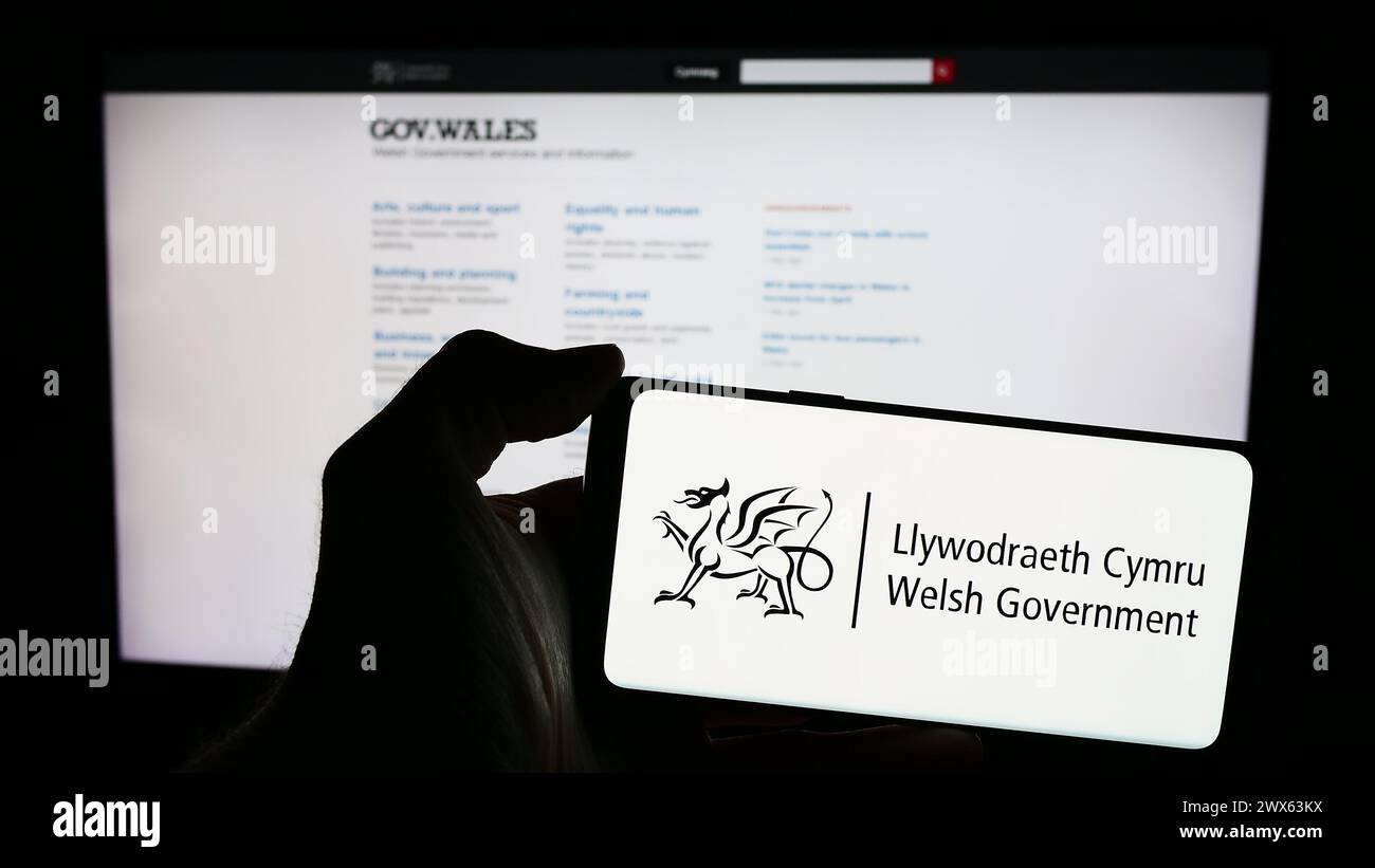 Person, die ein Mobiltelefon mit dem Logo der walisischen Regierung (Llywodraeth Cymru) vor der Webseite hält. Schwerpunkt auf dem Display des Telefons. Stockfoto