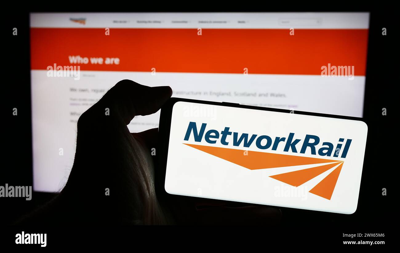 Person, die ein Mobiltelefon mit dem Logo des Eisenbahninfrastrukturunternehmens Network Rail Limited vor der Webseite hält. Schwerpunkt auf dem Display des Telefons. Stockfoto