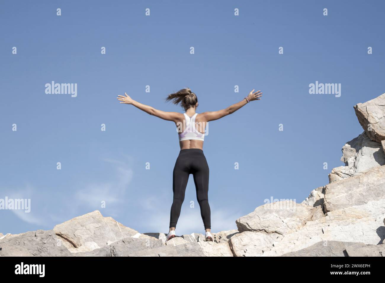 Eine Person steht triumphierend auf felsigem Gelände, mit ausgestreckten Armen, unter einem klaren blauen Himmel, und verkörpert Freiheit und Leistung Stockfoto