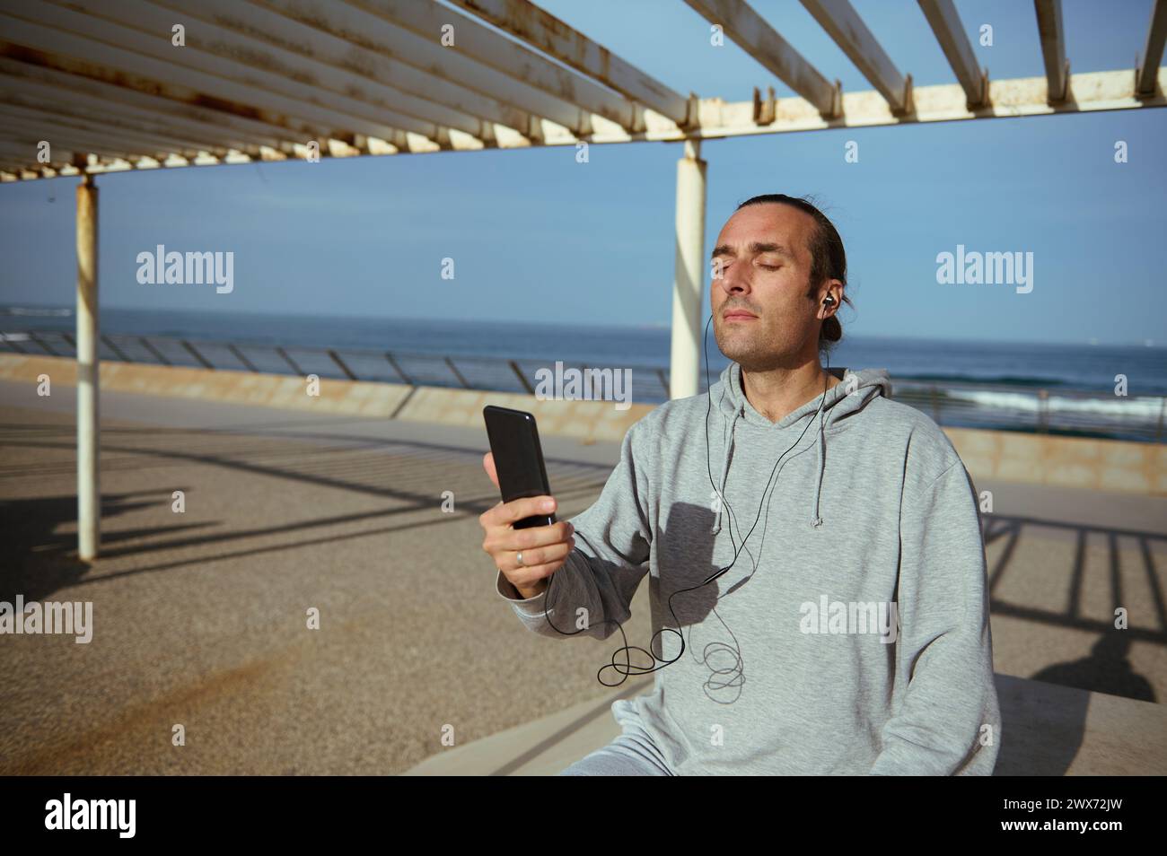 Junger kaukasischer Mann in grauer Sportbekleidung, mit Ohrhörern, Smartphone in der Hand, Musik hören, während er nach dem Workout ruht, auf einer Straße sitzt Stockfoto