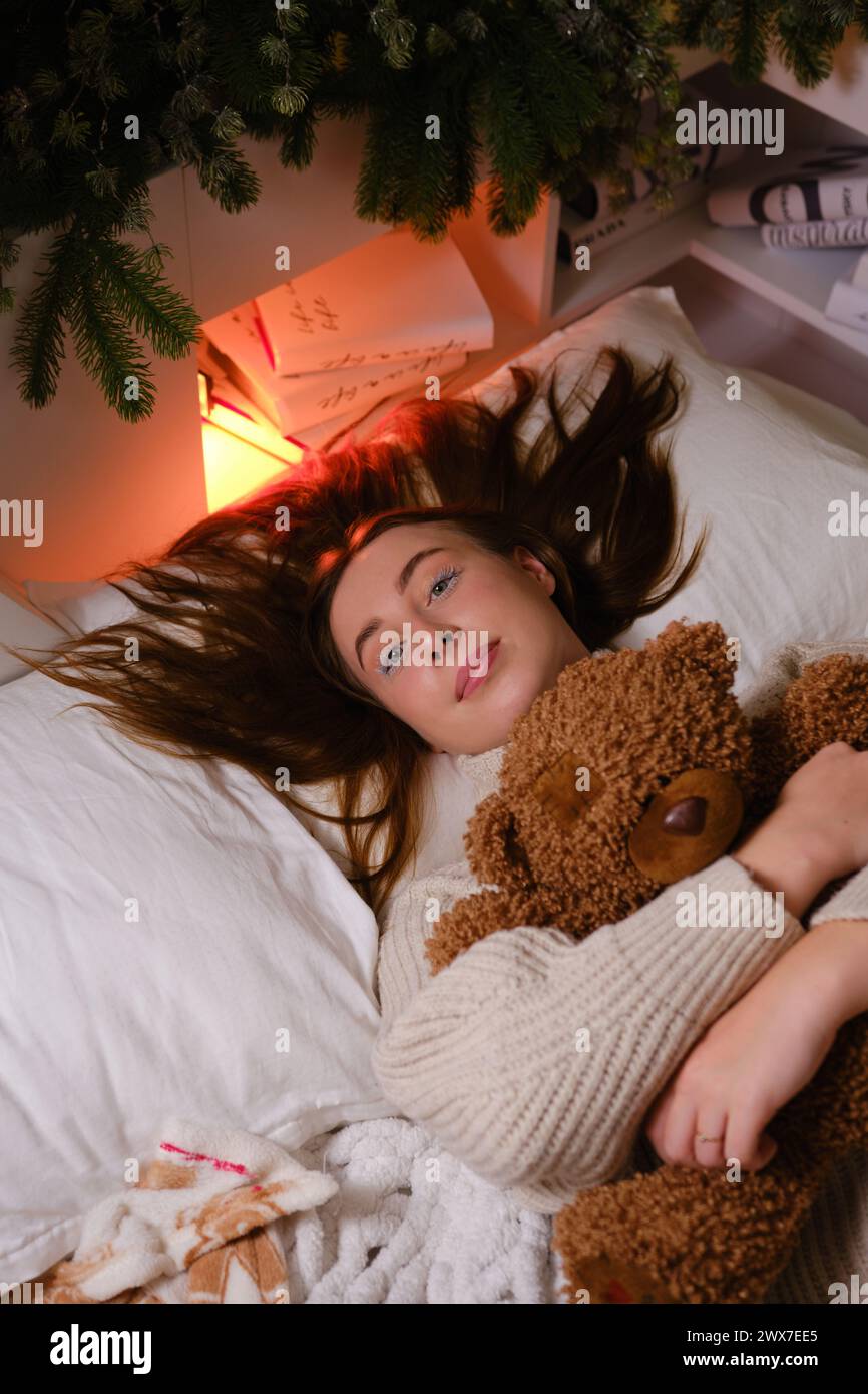 Fröhliche hübsche Frau in weißem Pullover mit braunem Teddybär-Spielzeug liegt auf Kissen unter dem Hintergrund des Weihnachtsbaums Stockfoto