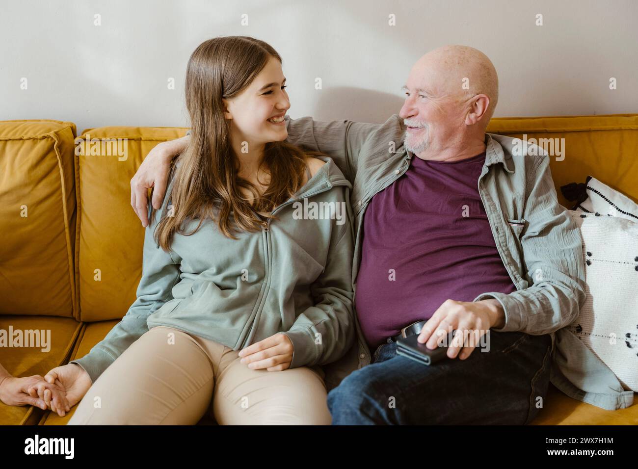 Ein lächelnder Seniorenmann sitzt mit Arm um die Teenager-Enkelin auf dem Sofa zu Hause Stockfoto