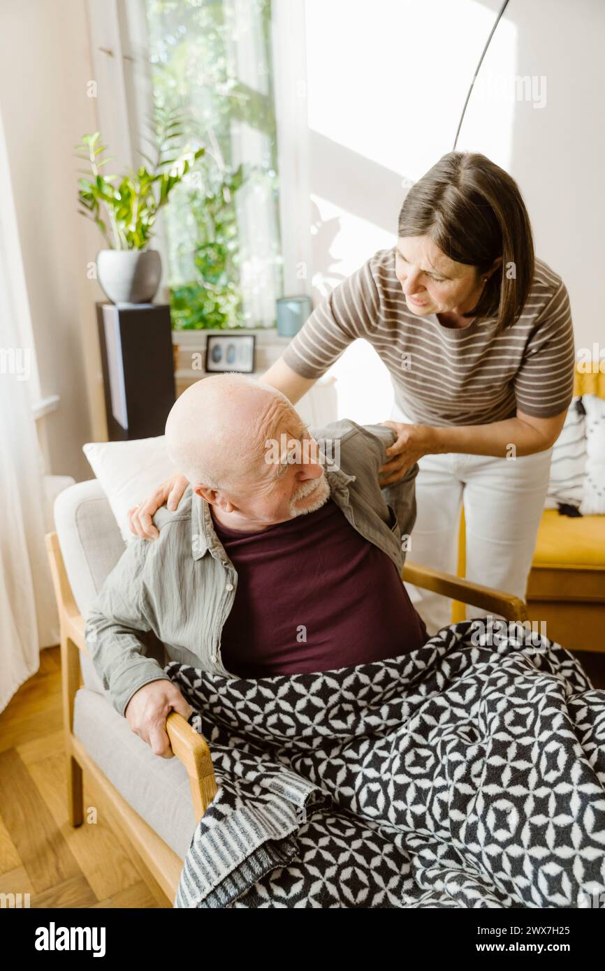 Reife Frau kümmert sich um einen älteren Mann, der zu Hause auf einem Stuhl sitzt Stockfoto