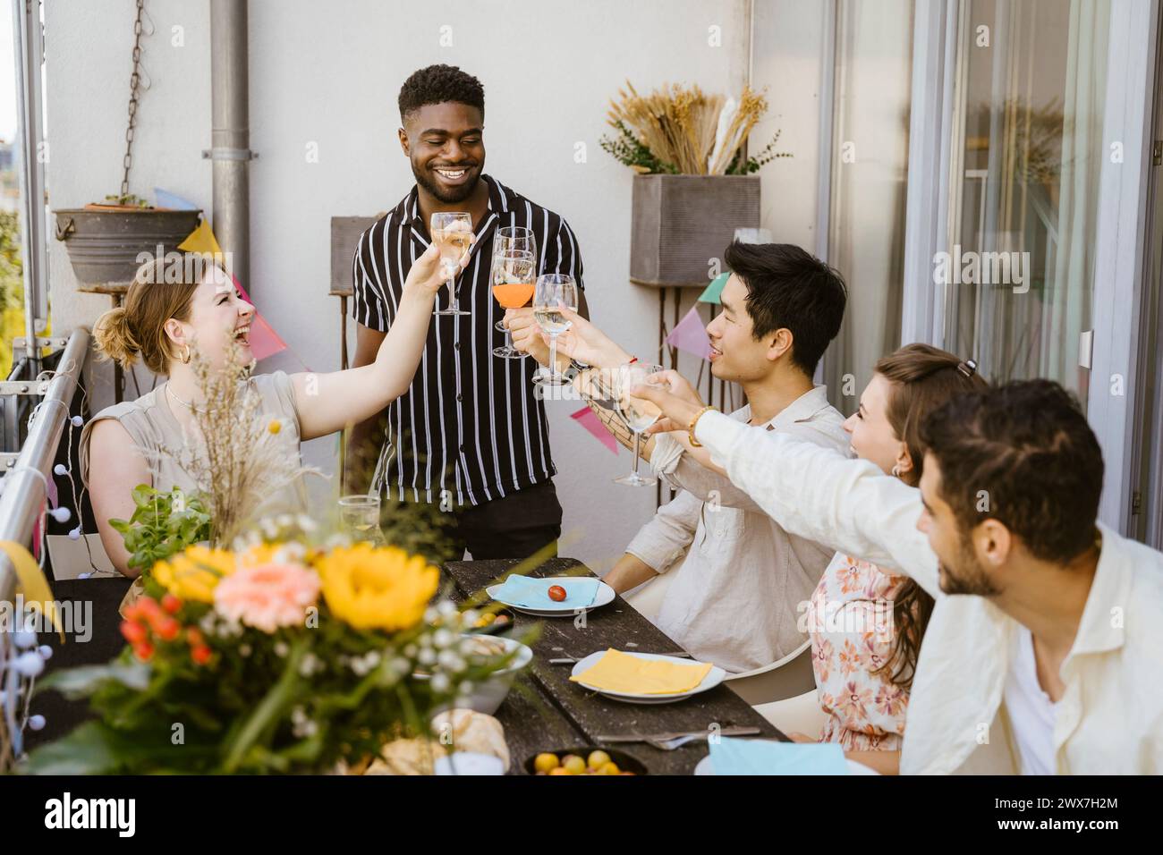Ein lächelnder junger Mann, der mit einer Gruppe männlicher und weiblicher Freunde während der Abendparty auf dem Balkon Getränke trinkt Stockfoto
