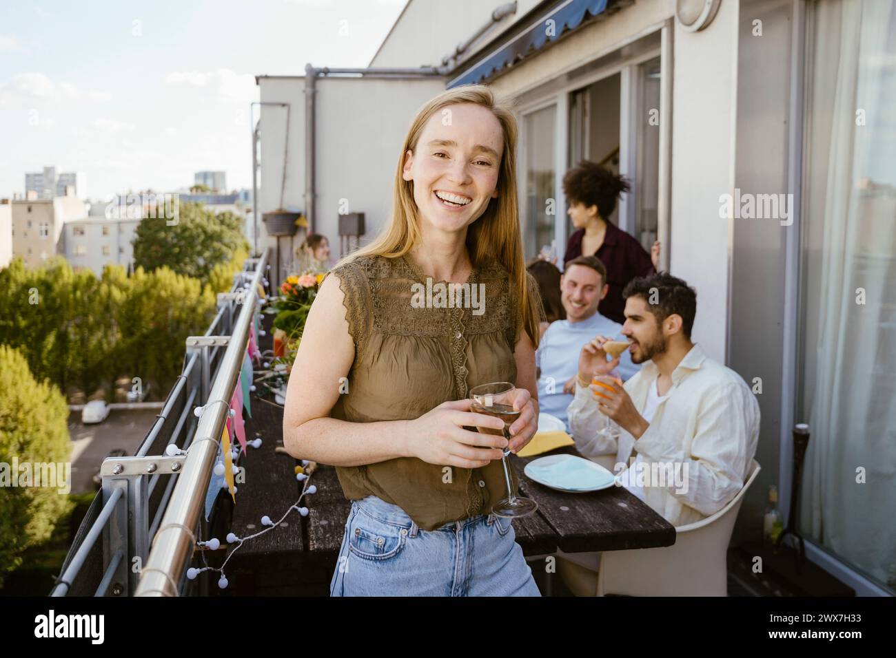 Porträt einer lächelnden Frau, die ein Glas in der Hand hält, während sie mit Freunden auf dem Balkon ein Abendessen feiert Stockfoto