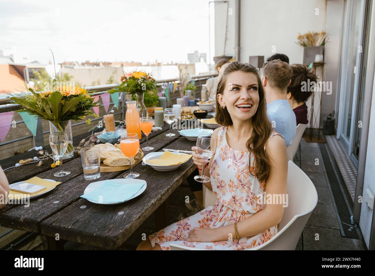 Lächelnde Frau, die Weinglas hält, während sie mit Freunden auf dem Stuhl sitzt, bei der Dinnerparty auf dem Balkon Stockfoto