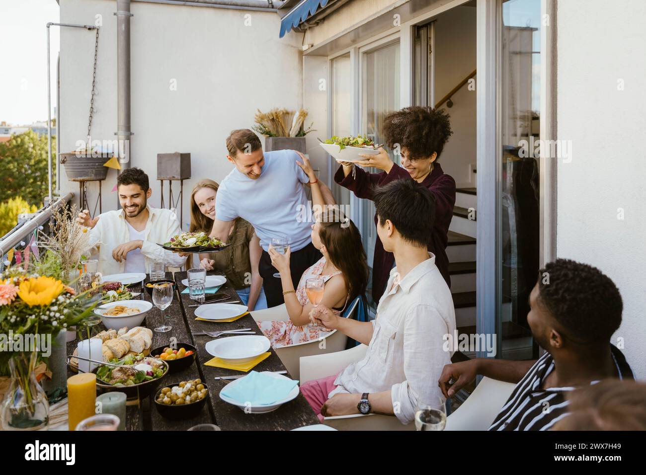 Glücklicher Mann und Frau servieren Essen für eine Gruppe von Freunden, die am Esstisch sitzen, während der Dinnerparty auf dem Balkon Stockfoto