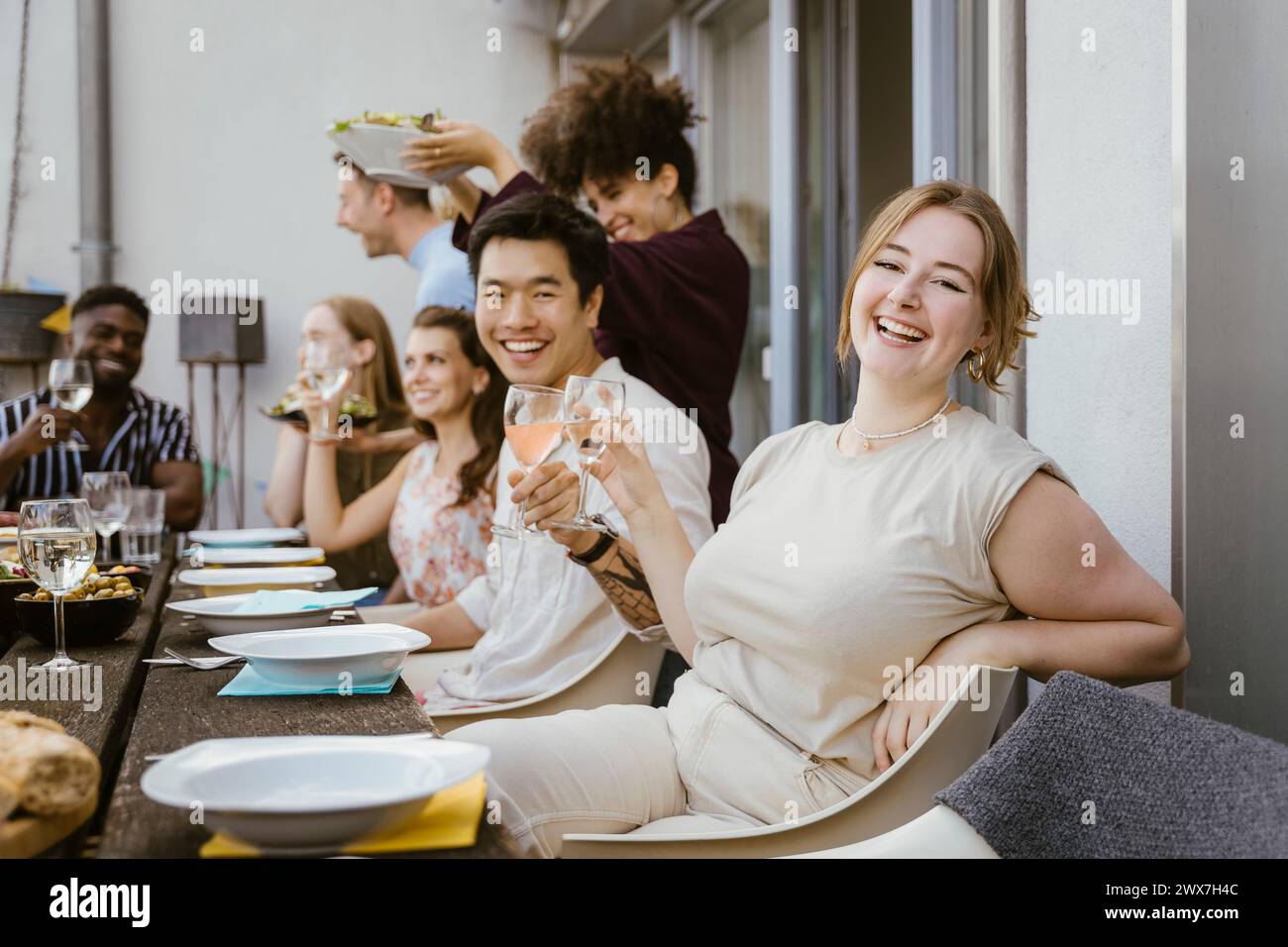 Porträt einer glücklichen jungen Frau, die Weinglas hält, während sie mit einer Gruppe von Freunden auf der Abendparty auf dem Balkon sitzt Stockfoto