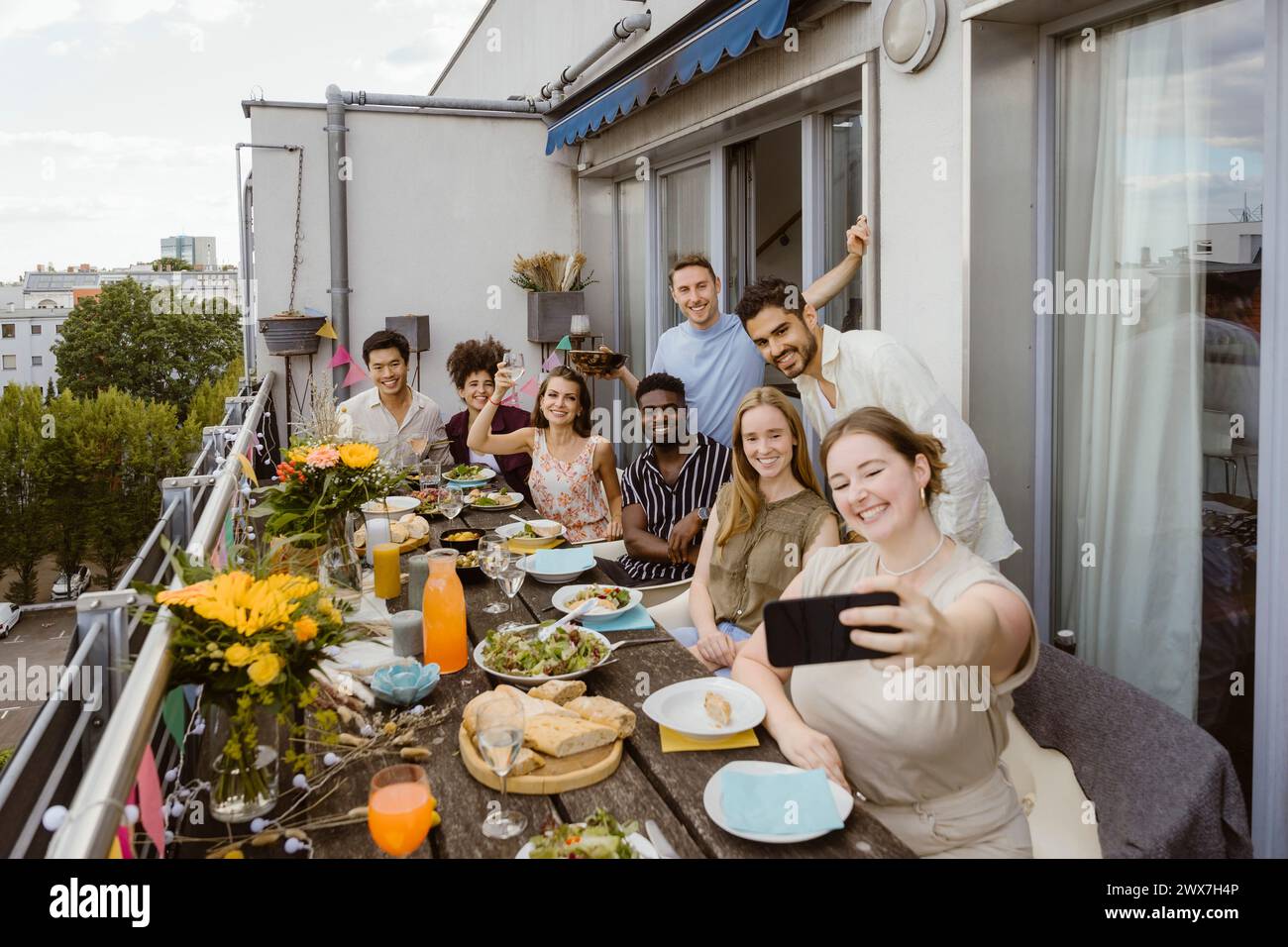 Lächelnde Frau macht Selfie mit einer Gruppe von Freunden über das Smartphone, während sie auf dem Balkon eine Dinnerparty feiert Stockfoto
