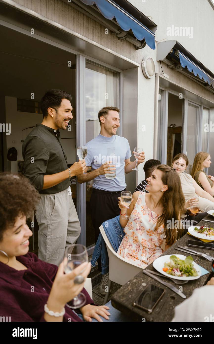 Lächelnde männliche Freunde, die mit einer Gruppe von Freunden bei der Dinnerparty auf dem Balkon Getränke genießen Stockfoto