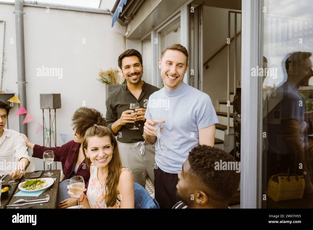 Glückliche männliche Freunde mit einer Gruppe von Freunden, die Getränke während der Dinnerparty auf dem Balkon genießen Stockfoto